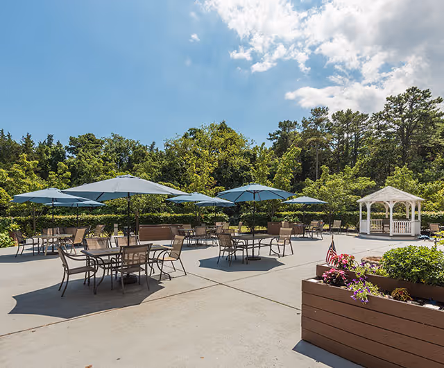 Outdoor patio with tables and blue umbrellas, planter boxes with flowers, and a white gazebo in front of trees under a sunny sky.