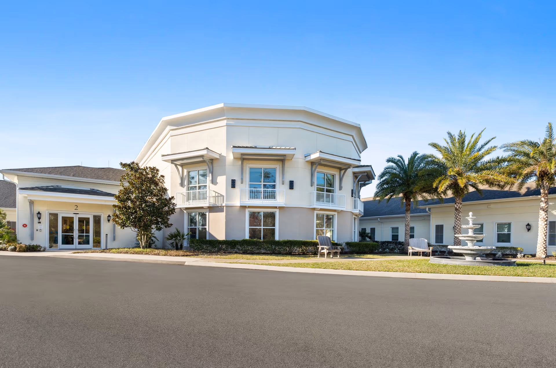 Exterior view of a senior living facility building with a light-colored facade, multiple windows, and a main entrance marked with the number 2. The area in front features a paved driveway, landscaped greenery, palm trees, benches, and a decorative water fountain under a clear blue sky.