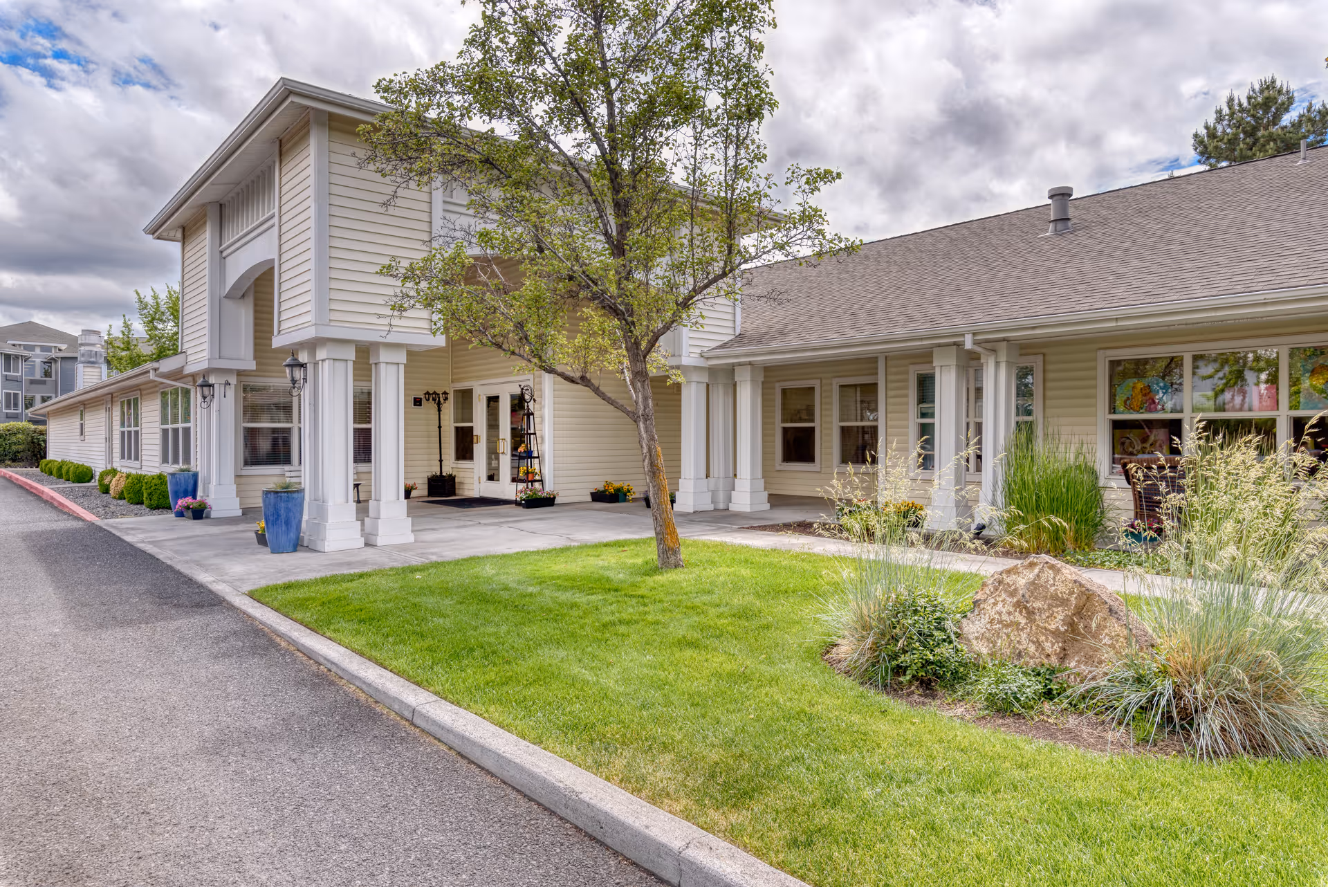 Exterior view of Awbrey Place Assisted Living and Memory Care building with a covered entrance, beige siding, white columns, a tree, green lawn, and landscaping with bushes and rocks under a cloudy sky.