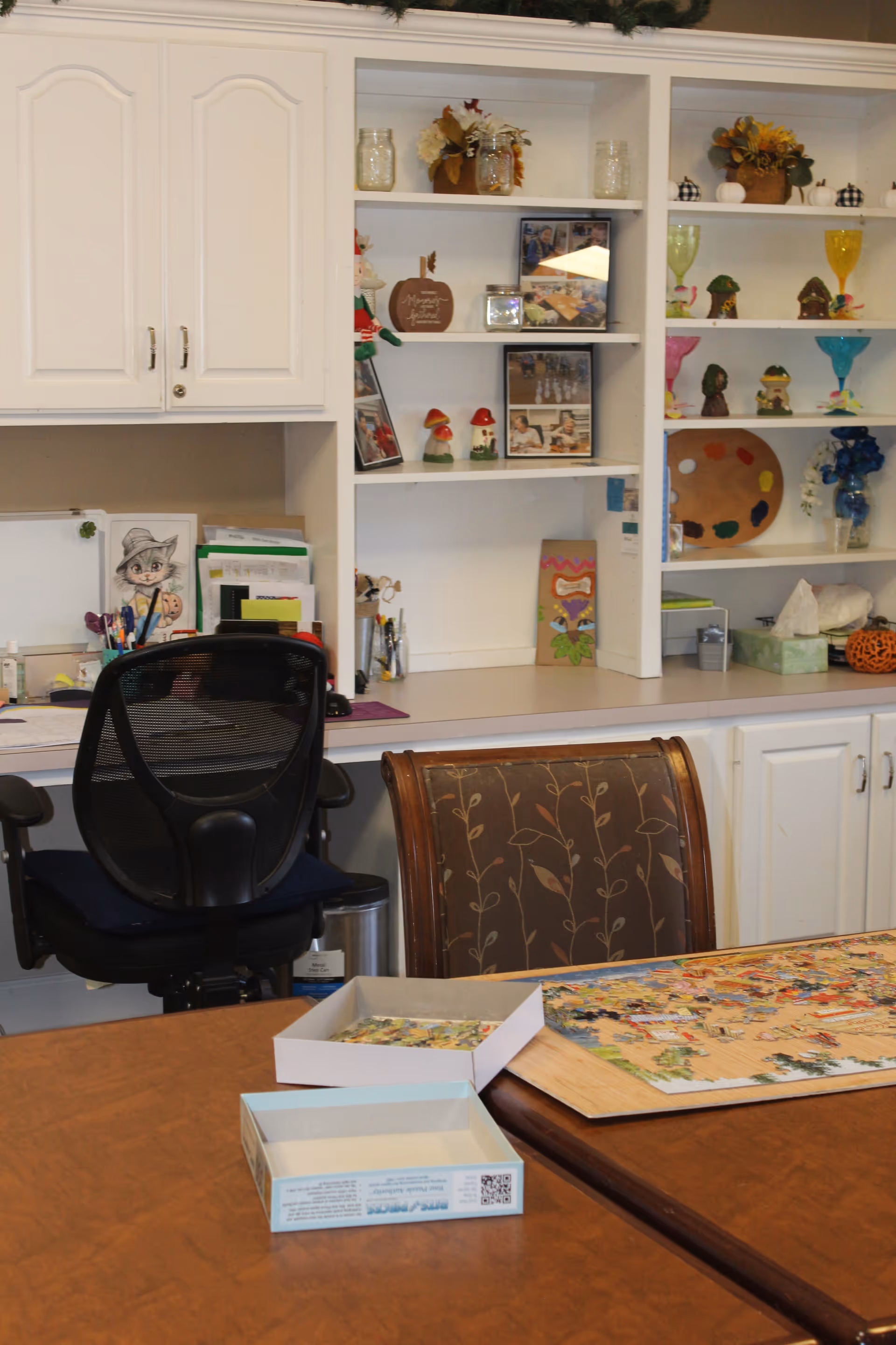 Community activity room with a table holding a partially completed puzzle, chairs, and built-in shelves and cabinets with decorative items.