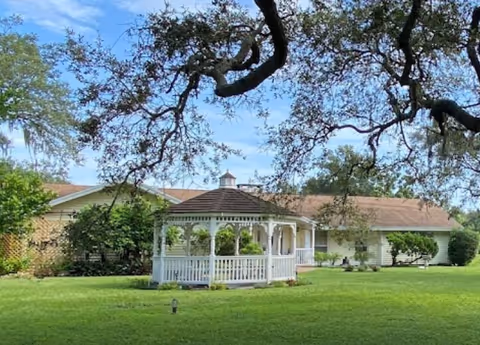 A white wooden gazebo with a brown shingled roof sits on a well-maintained green lawn in front of a single-story building with a light-colored exterior. Large tree branches with sparse leaves frame the top of the image under a blue sky with some clouds.