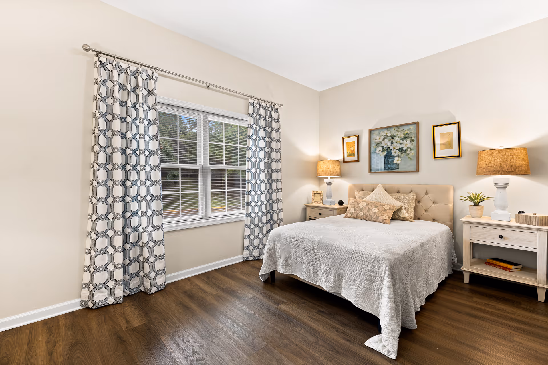 A neatly staged bedroom with a tufted bed, two bedside tables and lamps, patterned curtains over a window, and hardwood floors.