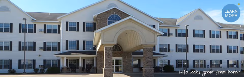 Front exterior view of a three-story senior living facility building with white siding, black window shutters, and a covered entrance supported by stone pillars. There are several windows and a clear blue sky above.
