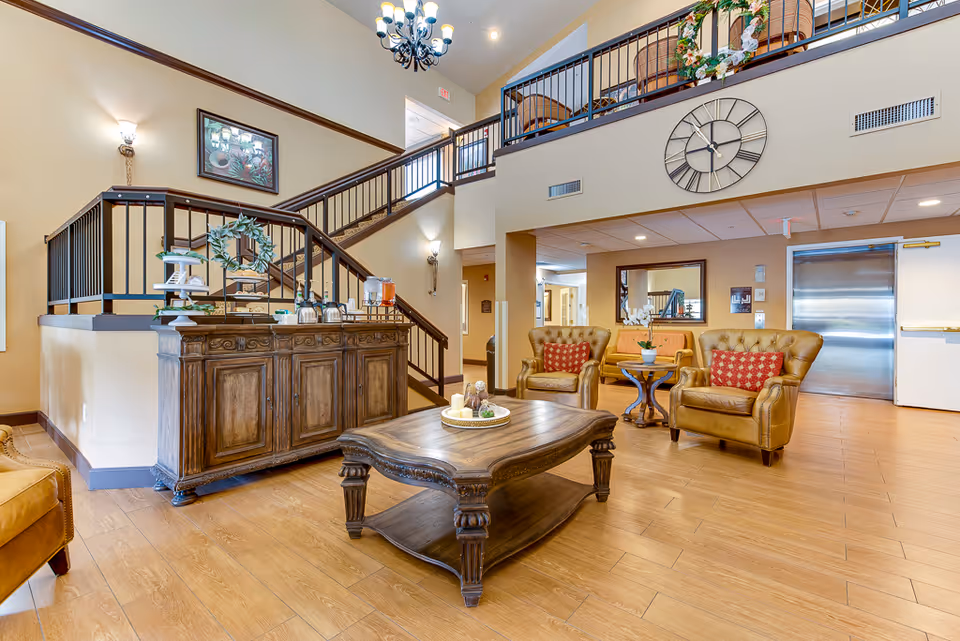 A bright and spacious senior living facility common area with wooden flooring, a large wooden coffee table with decorative items, two tan leather armchairs with red patterned cushions, a small round side table with a potted orchid, and a wooden cabinet with refreshments. A staircase with black railings leads to an upper level with additional seating. A large wall clock and framed artwork decorate the walls.