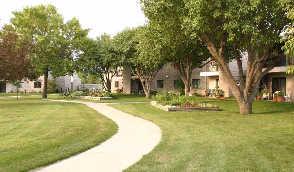A winding concrete pathway through a grassy lawn with trees and flower beds, leading to a multi-unit residential building with balconies and patios, surrounded by greenery.