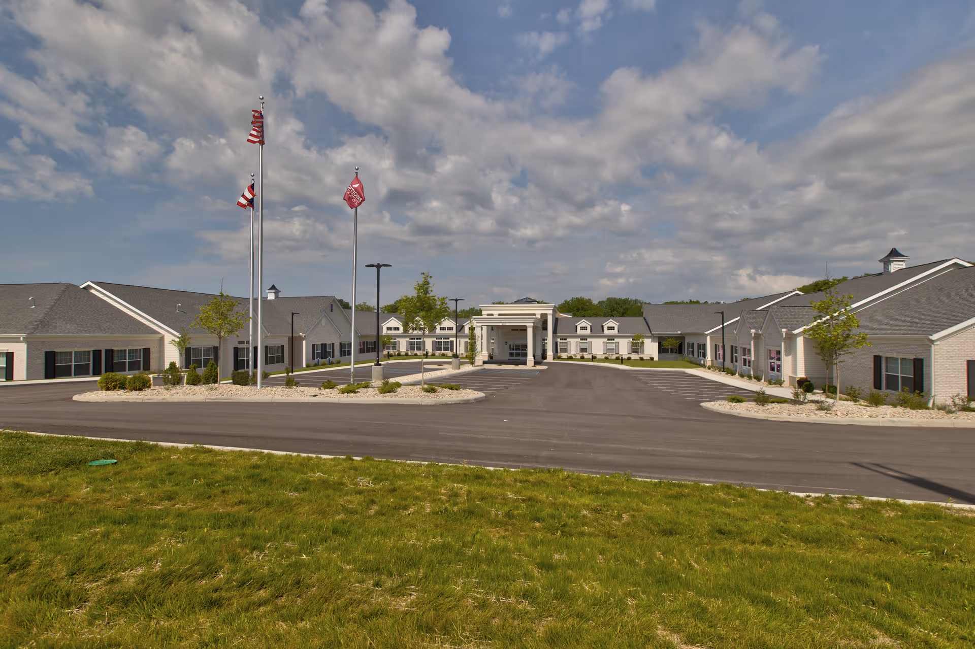 Front exterior view of Traditions of Lebanon senior living facility with a large driveway, three flagpoles with flags, manicured landscaping, and a partly cloudy sky.
