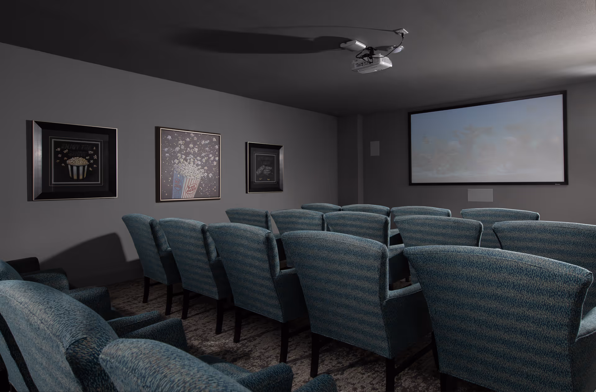 A small movie theater room with several rows of blue upholstered chairs facing a large projection screen. The walls are gray and decorated with framed pictures of popcorn and movie-related themes. A ceiling-mounted projector is visible above the seating area.