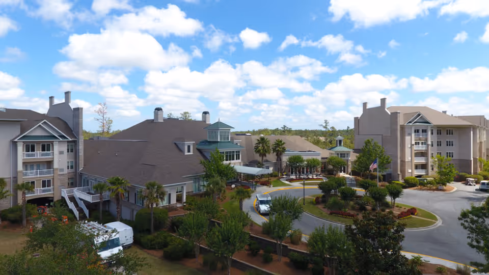Aerial view of Marshes of Skidaway Island senior living facility showing multiple connected buildings with beige and gray exteriors, surrounded by landscaped greenery, palm trees, and a circular driveway with a vehicle. The sky is blue with scattered clouds.