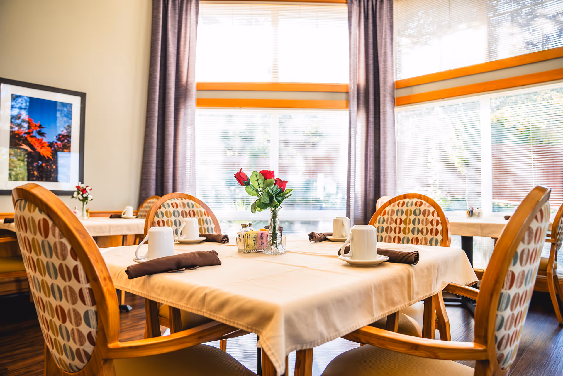 Sunlit dining room with a set table featuring a vase of red roses, cups, napkins, and patterned wooden chairs.