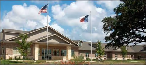 Exterior view of a senior living facility with flags, entrance, and landscaping.