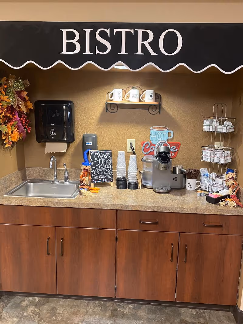 A coffee station with a sink, paper towel dispenser, coffee maker, disposable cups, coffee creamers, and utensils. Above the counter is a black awning with the word 'BISTRO' in white letters. There is a decorative fall wreath on the left wall and a small shelf with mugs on the back wall.