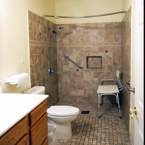 A bathroom with a tiled walk-in shower area featuring a handheld showerhead, grab bars, and a shower chair. The bathroom also includes a toilet, a wooden vanity with a white countertop, and a toilet paper holder mounted on the wall.