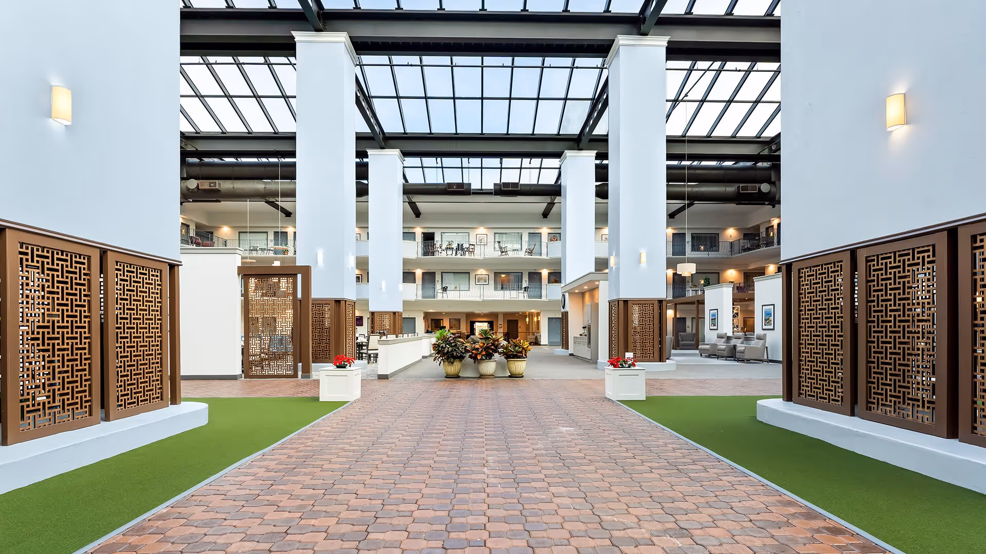 Spacious and bright interior atrium of a senior living facility with a high glass ceiling, white pillars, decorative wooden panels, potted plants, and seating areas visible in the background.