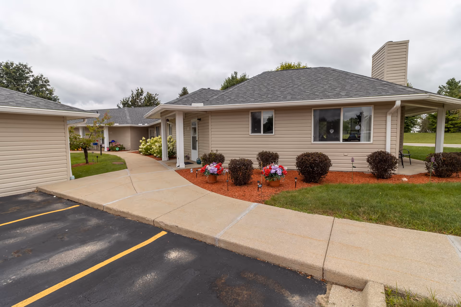 Exterior view of a single-story beige building with a gray roof at Independence Village of Saginaw. The building is surrounded by a paved walkway, green grass, and landscaped bushes with colorful flowers. There is a parking area with marked spaces in the foreground under a cloudy sky.