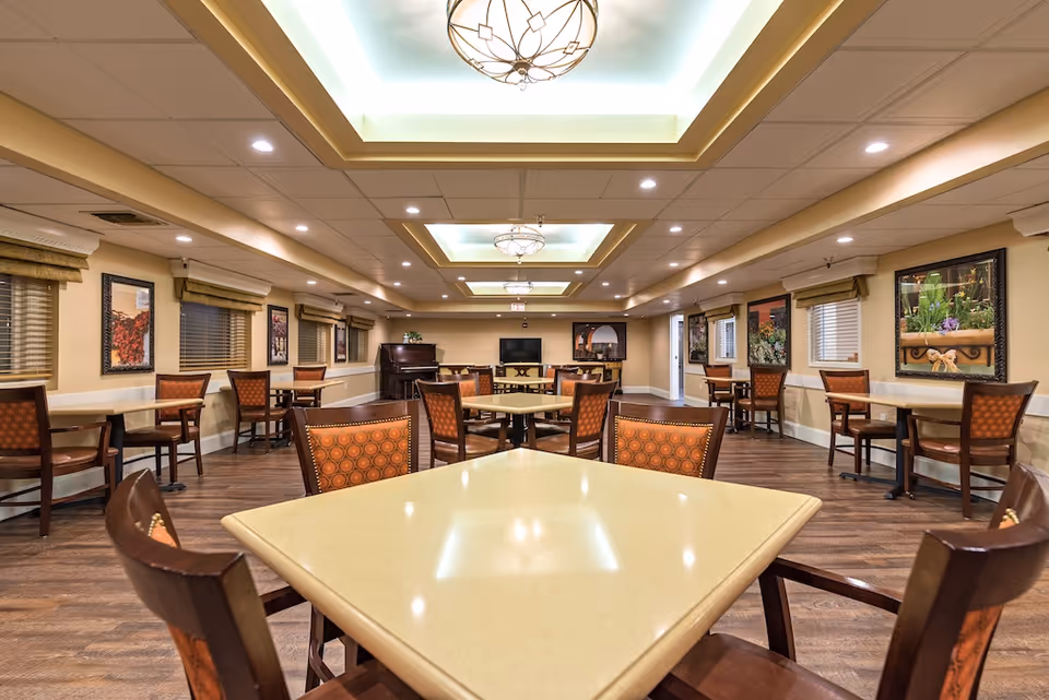 A spacious dining room with multiple tables and chairs arranged neatly. The room features warm lighting with decorative ceiling fixtures, framed artwork on the walls, and windows with blinds and valances. The floor is wooden, and there is a piano and a television at the far end of the room.
