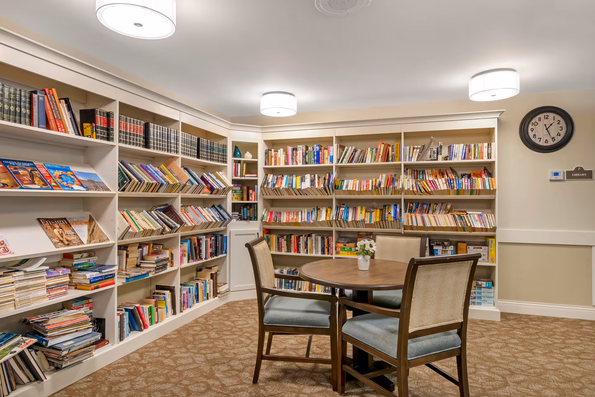 A cozy library room with white built-in bookshelves filled with books along two walls. In the center, there is a round wooden table with four cushioned chairs. The room is well-lit with ceiling lights, and a clock is mounted on the wall above a sign that reads 'Library'.