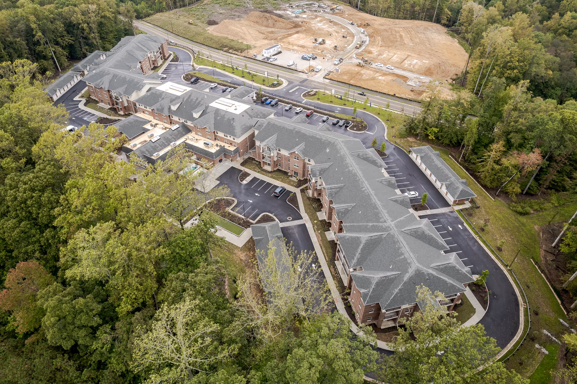 Aerial view of Virginia Greens Retirement Community showing a large multi-wing building surrounded by trees and parking lots, with a construction site visible in the background.