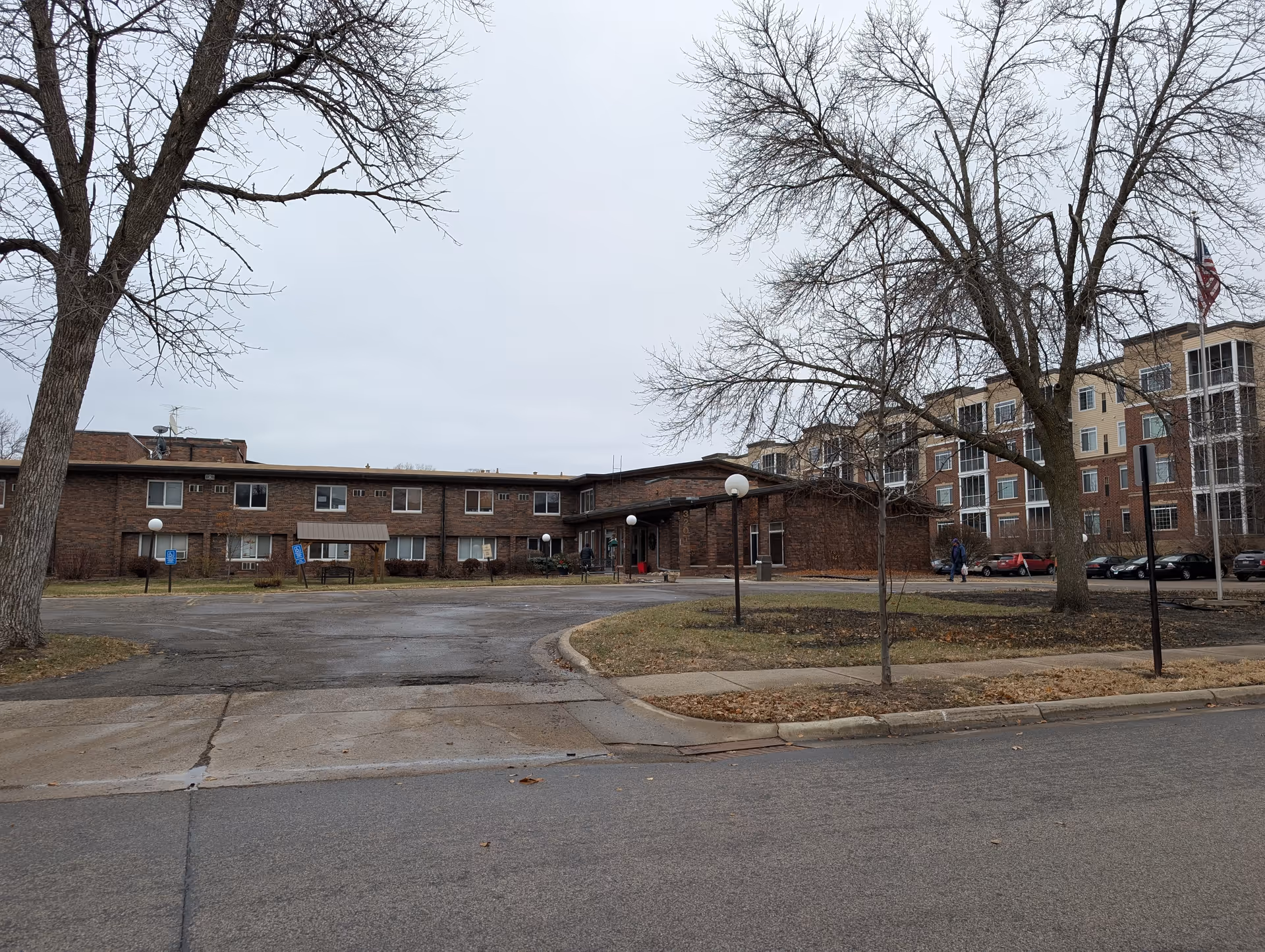 Exterior view of a senior living facility with a brick building and a parking area in front. Leafless trees are visible around the property, and an American flag is flying on a flagpole near the building. The sky is overcast.