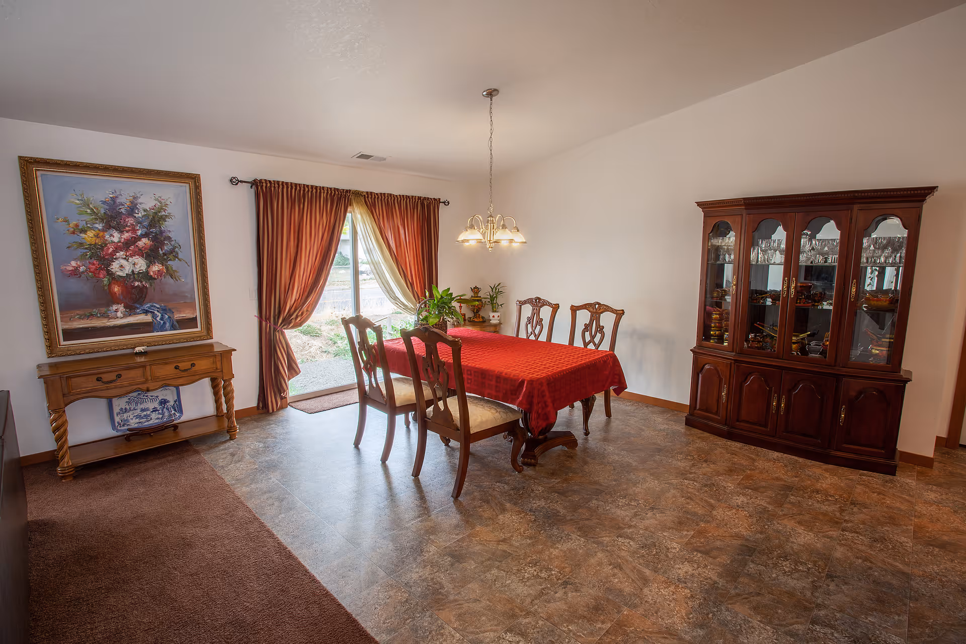 A dining room with a rectangular wooden table covered with a red tablecloth and six matching wooden chairs. There is a wooden china cabinet with glass doors displaying glassware on the right side. On the left side, there is a wooden console table with a large framed floral painting above it. The room has a sliding glass door with red curtains letting in natural light. The floor is a mix of carpet and tile.