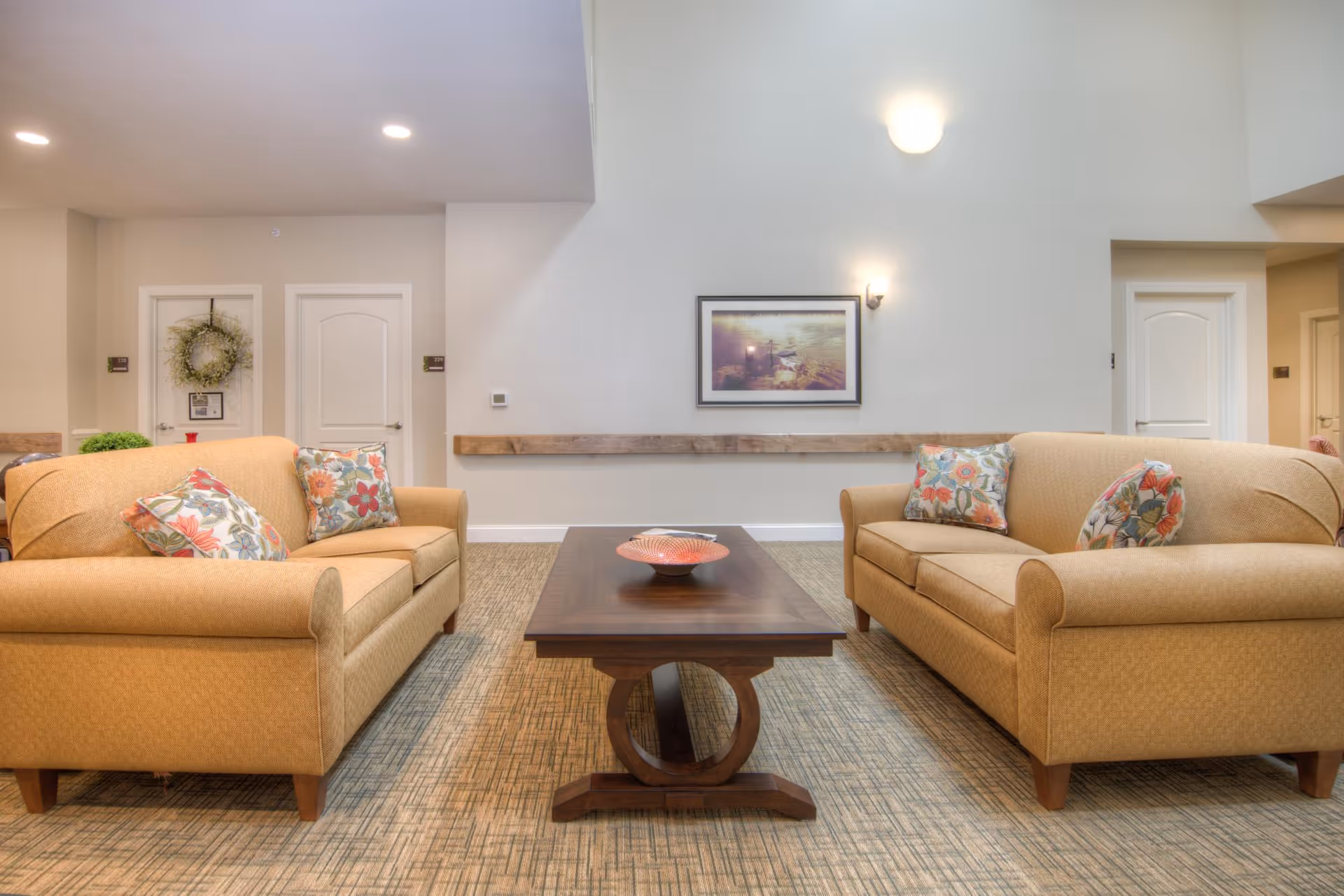 A cozy seating area in a senior living facility with two beige sofas facing each other, each adorned with floral patterned throw pillows. Between the sofas is a wooden coffee table with a decorative bowl. The room has light-colored walls, a framed picture, and soft lighting fixtures. There are two white doors in the background, one decorated with a wreath.