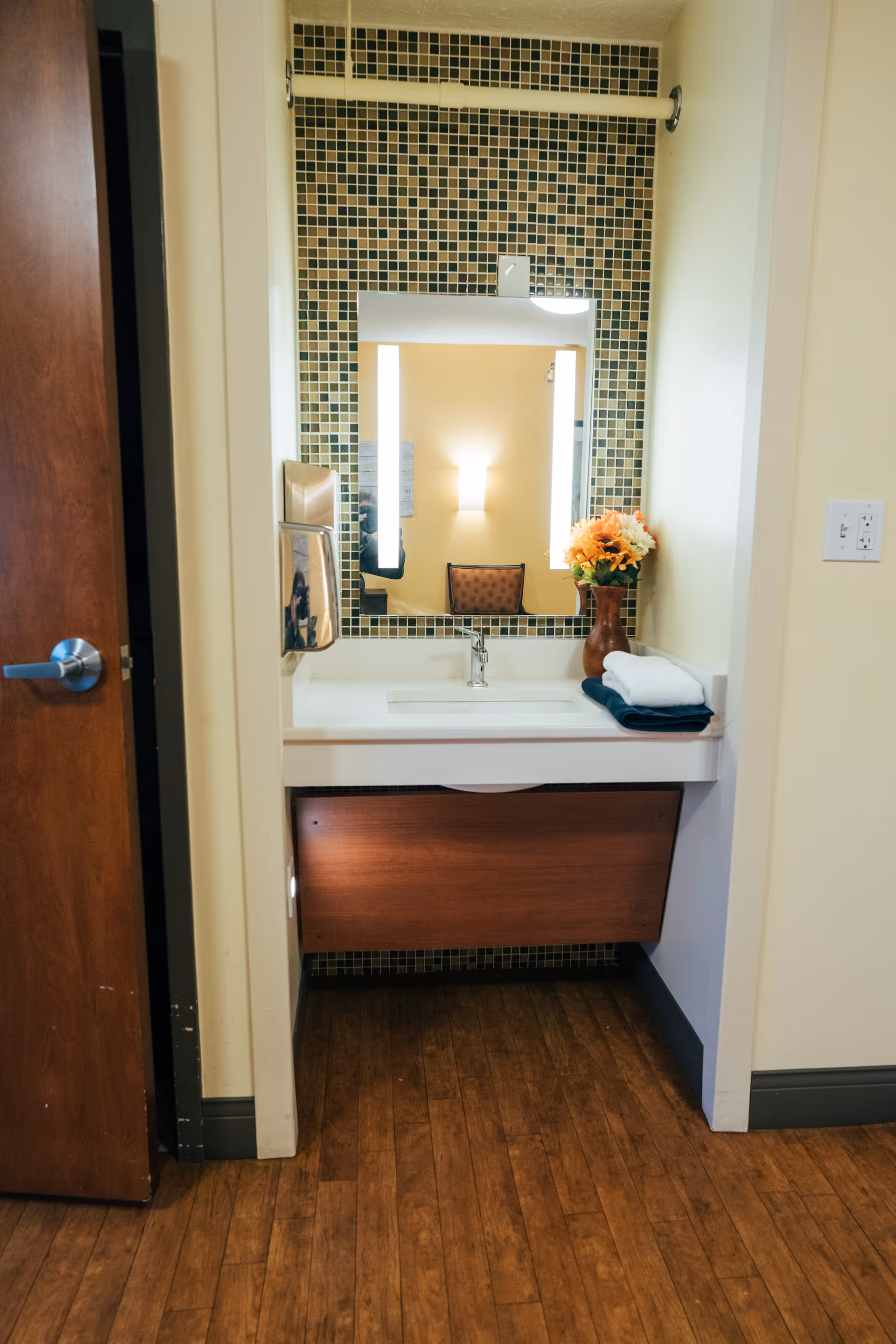 A small bathroom sink area with a rectangular mirror framed by small square mosaic tiles in shades of green and brown. The sink countertop holds a brown vase with orange and white flowers and neatly folded white and dark blue towels. To the left of the sink is a wall-mounted soap dispenser. The floor is wooden, and a partially open wooden door with a silver handle is visible on the left side.