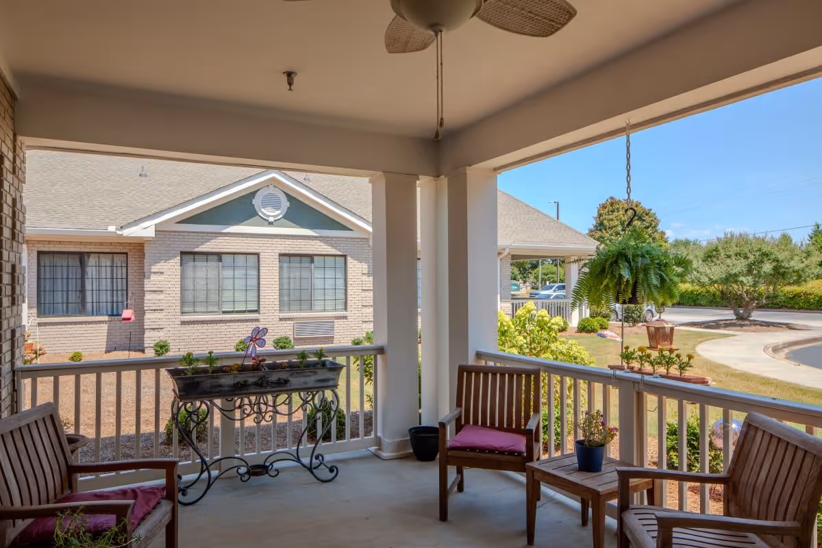 Covered outdoor patio area with wooden chairs and a small table, decorated with potted plants and a hanging fern. The patio overlooks a garden area with shrubs and a building in the background under a clear blue sky.