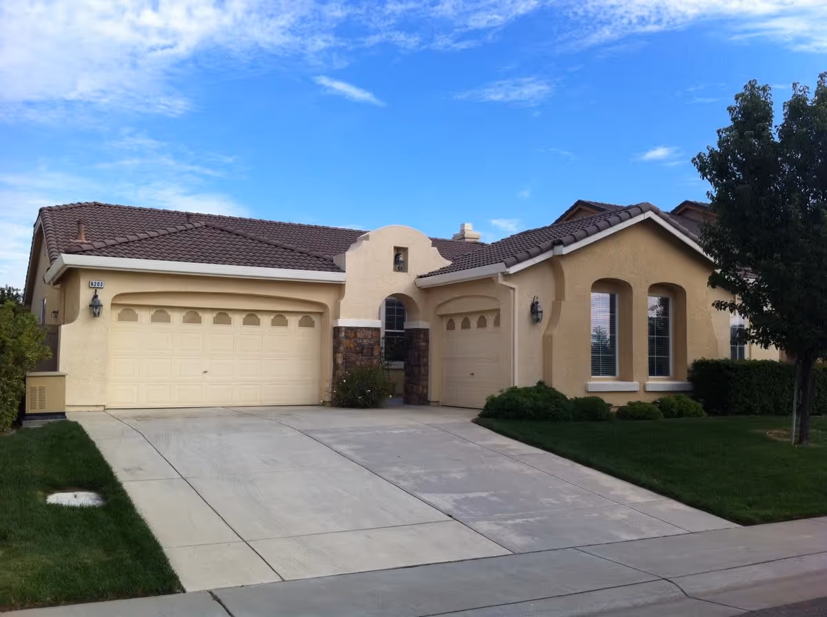 Exterior view of a single-story residential building with beige stucco walls, brown tiled roof, two garages, a driveway, green lawn, and a tree under a partly cloudy blue sky.