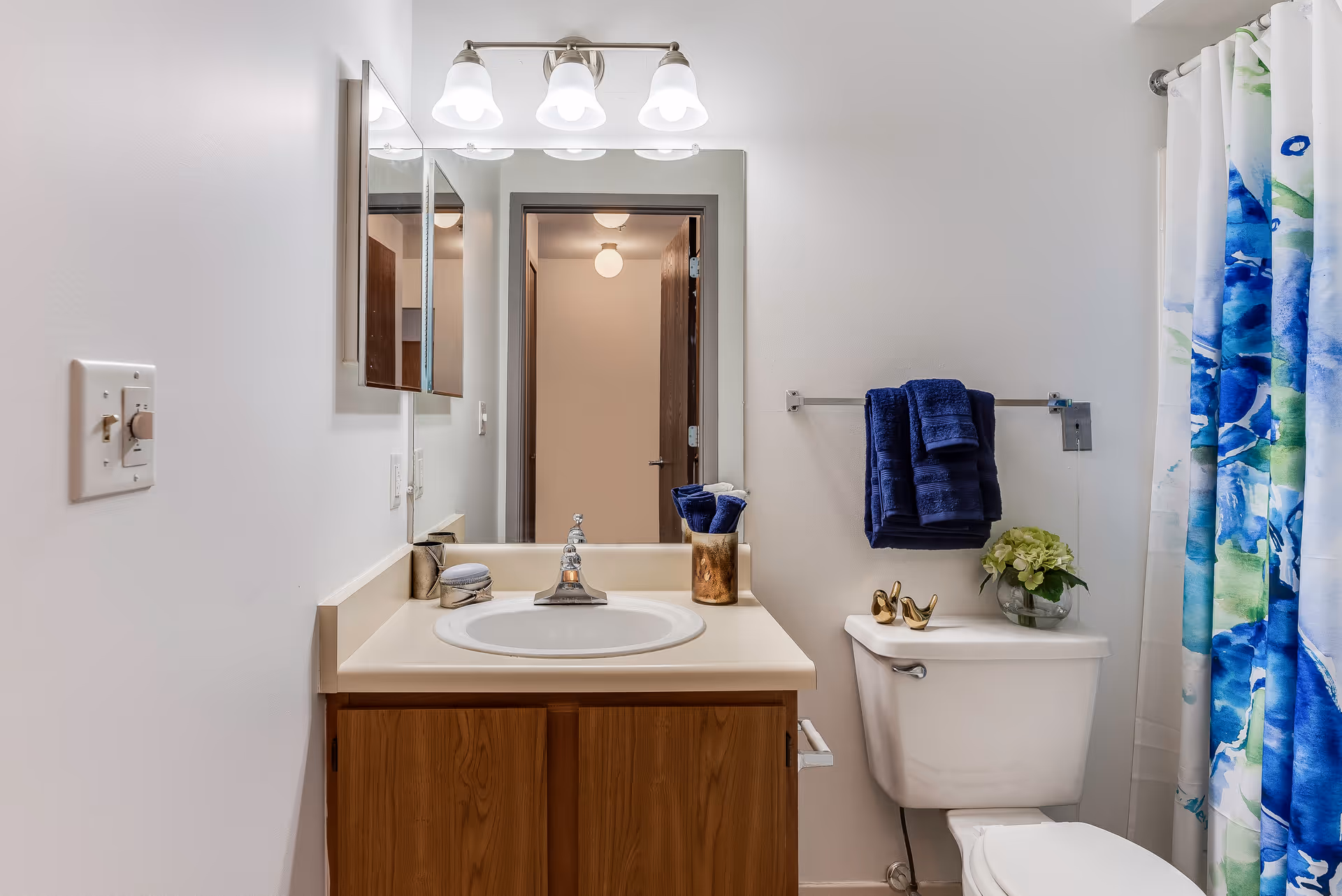 A clean bathroom with a wooden vanity cabinet, a white sink, and a mirror above it with three light fixtures. To the right, there is a toilet with a small vase of flowers on the tank and a towel rack holding dark blue towels. A colorful blue and green shower curtain is partially visible on the far right.