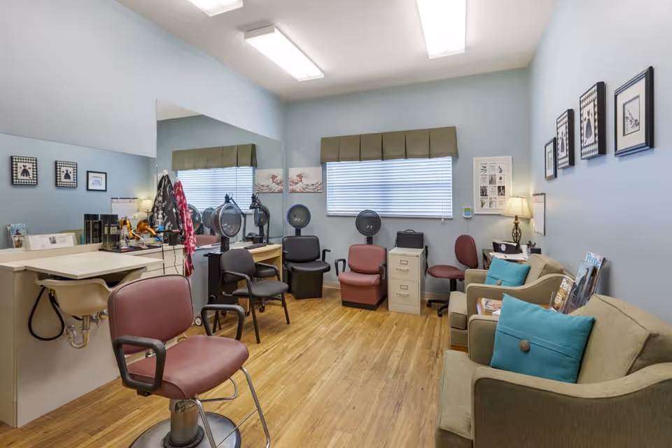 Interior of a salon room with multiple salon chairs, hair dryers, a large mirror on one wall, a sink, and a seating area with two green armchairs with blue pillows. The room has light blue walls, wooden flooring, and framed pictures on the walls.