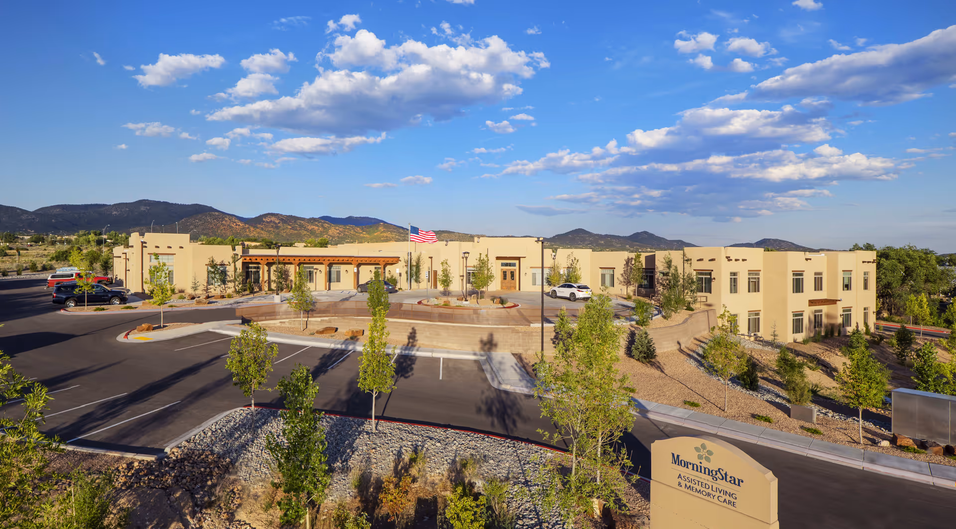 Exterior view of MorningStar Assisted Living & Memory Care of Santa Fe facility with a clear blue sky, mountains in the background, a parking lot with several cars, and landscaped greenery around the building.