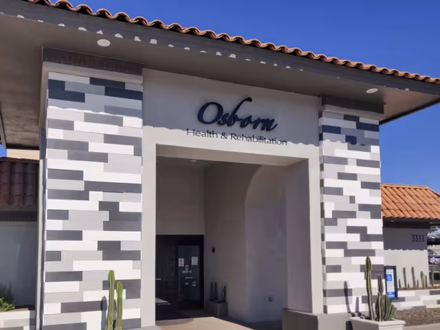 Entrance of Osborn Health & Rehabilitation building with a tiled roof and decorative gray and white brick columns on either side. There are a few tall cacti planted near the entrance under a clear blue sky.