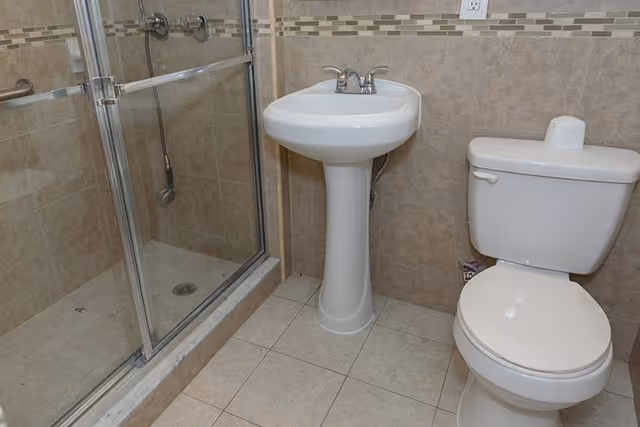 A clean bathroom featuring a glass-enclosed shower with chrome fixtures, a white pedestal sink with a chrome faucet, and a white toilet with a roll of toilet paper on top. The walls are tiled with beige tiles and a decorative tile border, and the floor is covered with light-colored tiles.
