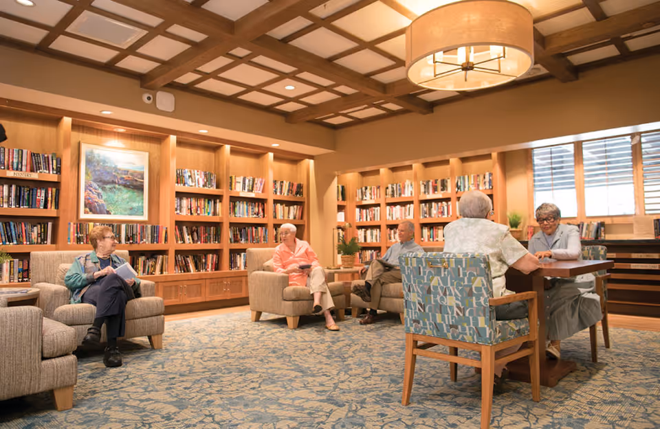 A cozy library room in a senior living facility with wooden bookshelves filled with books along the walls. Five elderly people are seated comfortably; two are sitting in armchairs reading, while two others are seated at a table engaged in conversation. The room features a patterned carpet, a large ceiling light fixture, and a painting on the wall.