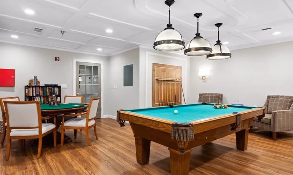 Community recreation room with a pool table, card table and chairs, bookshelves, and lounge seating under pendant lights.