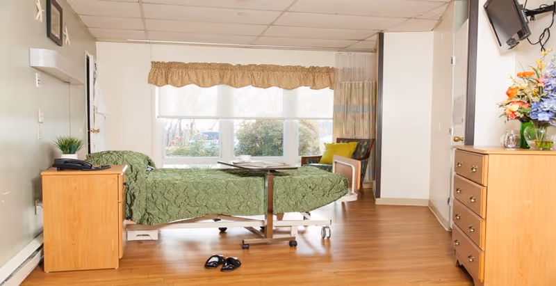 A senior living facility bedroom with a hospital-style bed covered in a green quilt, a wooden nightstand with a phone and a small plant, a wooden dresser with a vase of colorful flowers, a wall-mounted TV, a chair with a green cushion near a window with beige curtains, and wooden flooring.