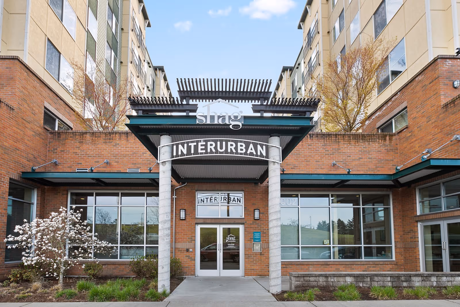 Front entrance of Interurban Senior Living facility with brick exterior, large windows, and a covered entryway supported by two columns. There are small landscaped areas with bushes and a flowering tree on either side of the entrance.