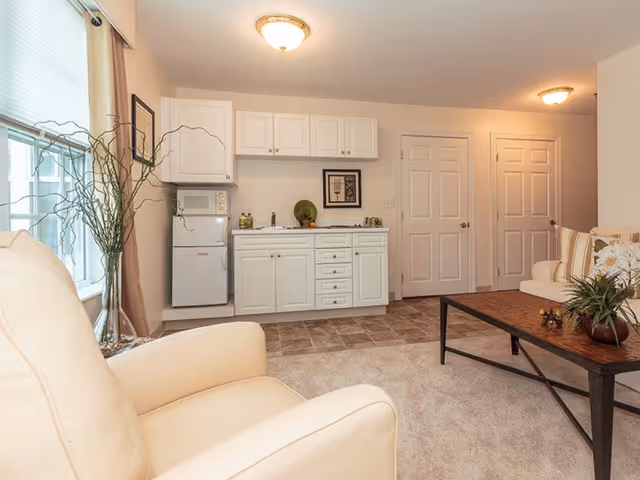 A cozy living area in Zelma Lacey House featuring a beige armchair and a sofa with floral cushions around a wooden coffee table. The room includes a small kitchenette with white cabinets, a mini refrigerator, a microwave, and a sink. Two closed white doors are visible in the background, and the space is softly lit by ceiling lights.