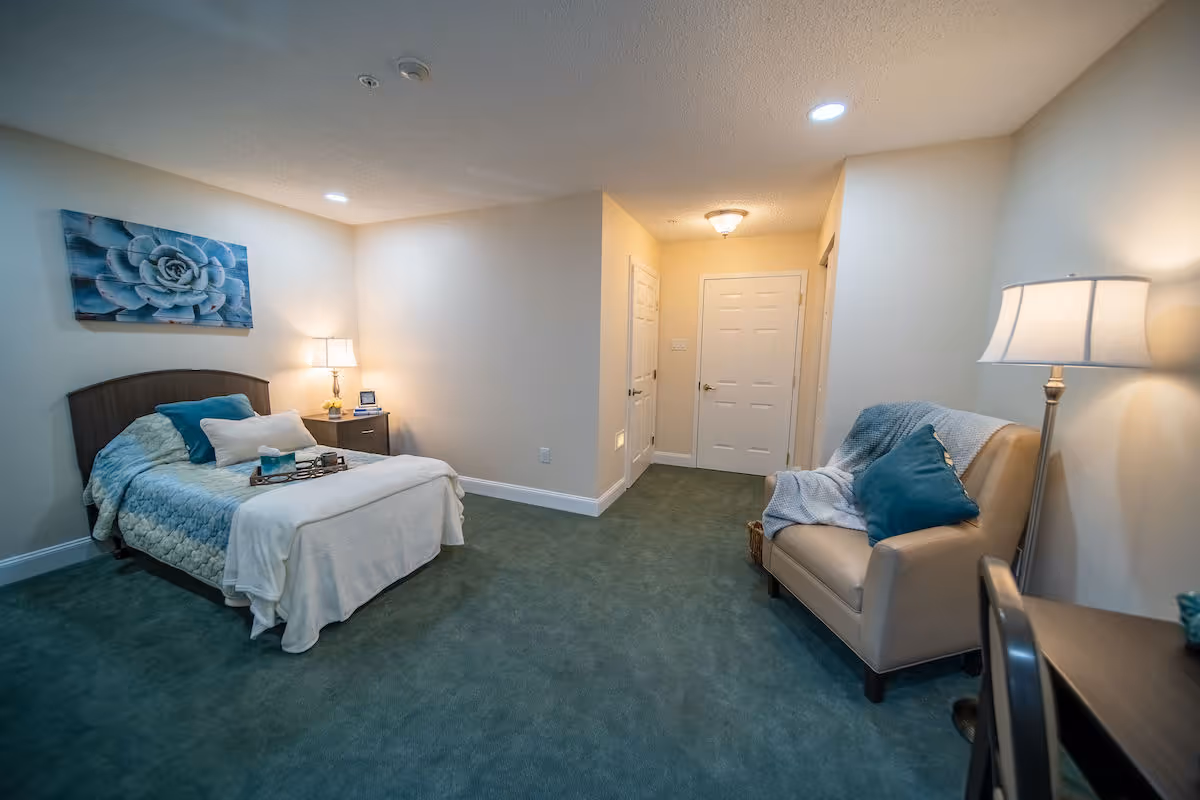 A cozy bedroom with a bed covered in blue and white bedding, a nightstand with a lamp and clock, a beige armchair with a blue pillow and throw blanket, and a floor lamp. The room has green carpet, light-colored walls, and a door leading to another area.