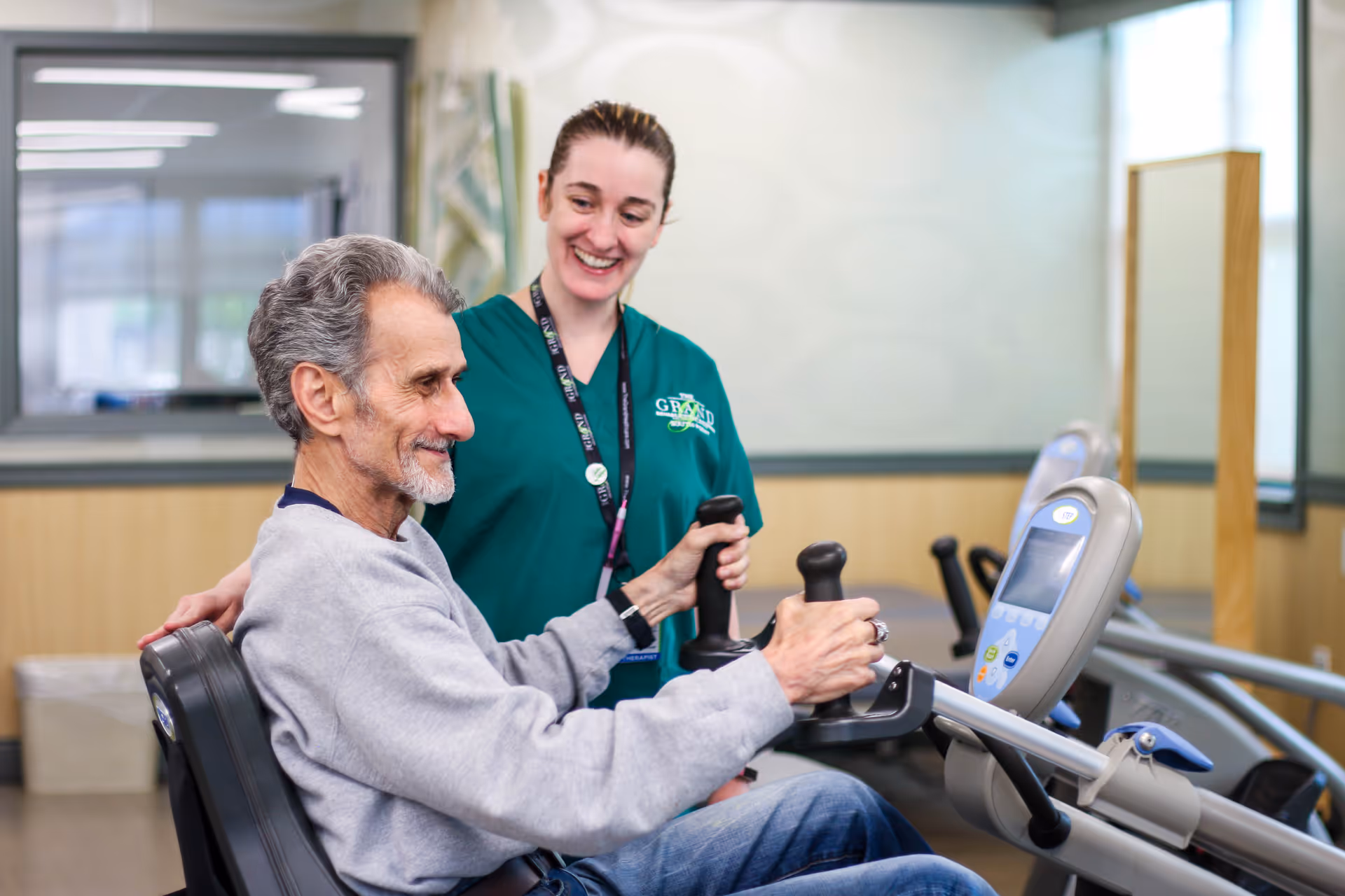 An elderly man exercises on a seated arm bike machine in a rehabilitation room while a female therapist in green scrubs smiles and assists him.