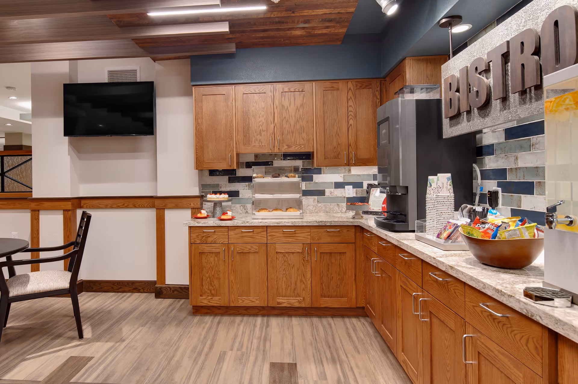 Interior view of a bistro area in a senior living facility with wooden cabinets, a countertop displaying snacks and desserts, a beverage dispenser, and a large sign reading 'BISTRO'. There is a flat-screen TV mounted on the wall and a round table with chairs nearby.