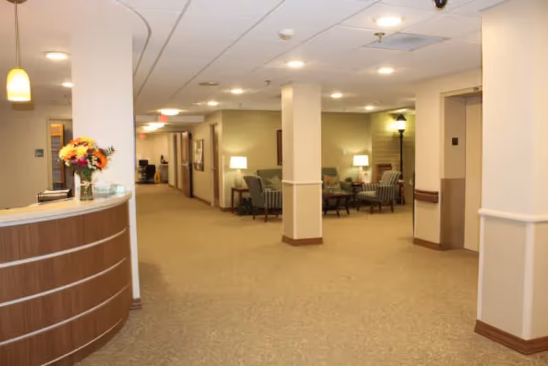 Interior view of a senior living facility hallway with a curved reception desk on the left adorned with a vase of flowers. The hallway leads to a seating area with several armchairs, side tables, and lamps. The walls and carpet are beige, and the ceiling has recessed lighting. An elevator is visible on the right side.