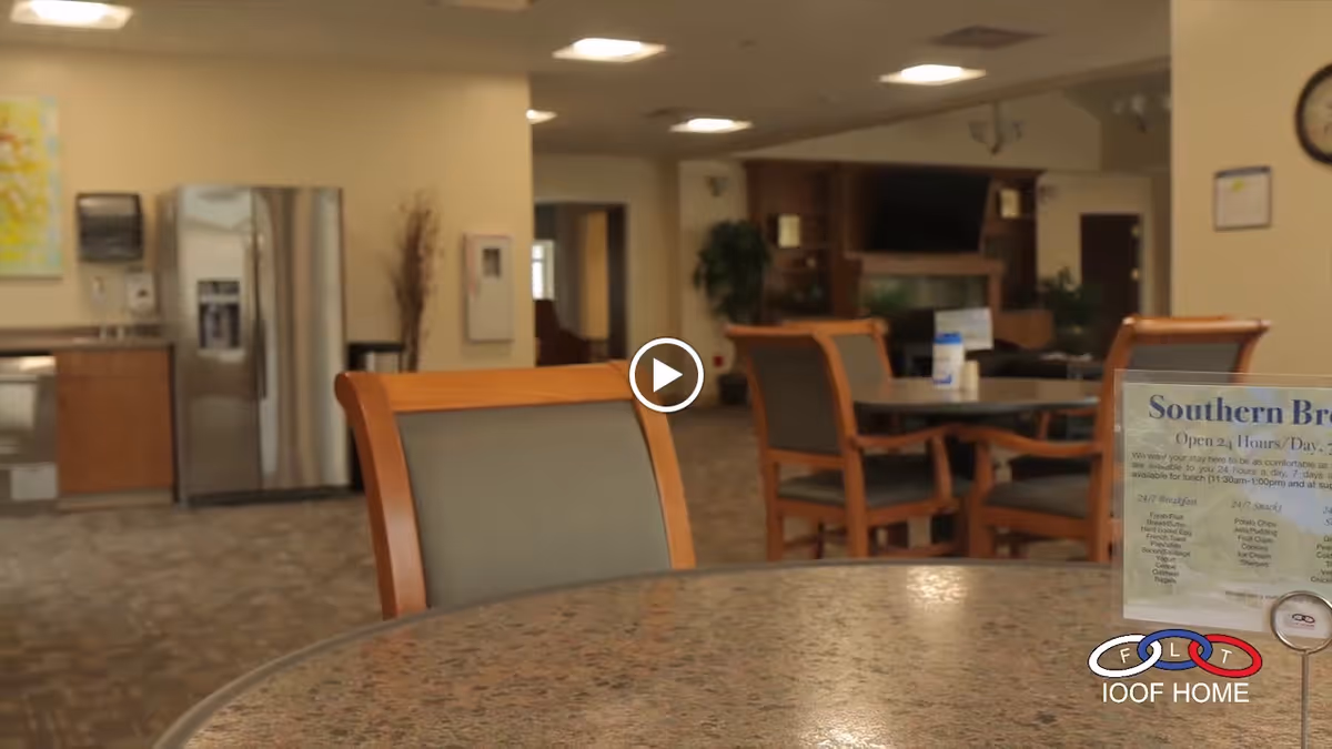 Round table and chairs in a senior living facility dining/lounge area with a kitchenette and menu sign visible.