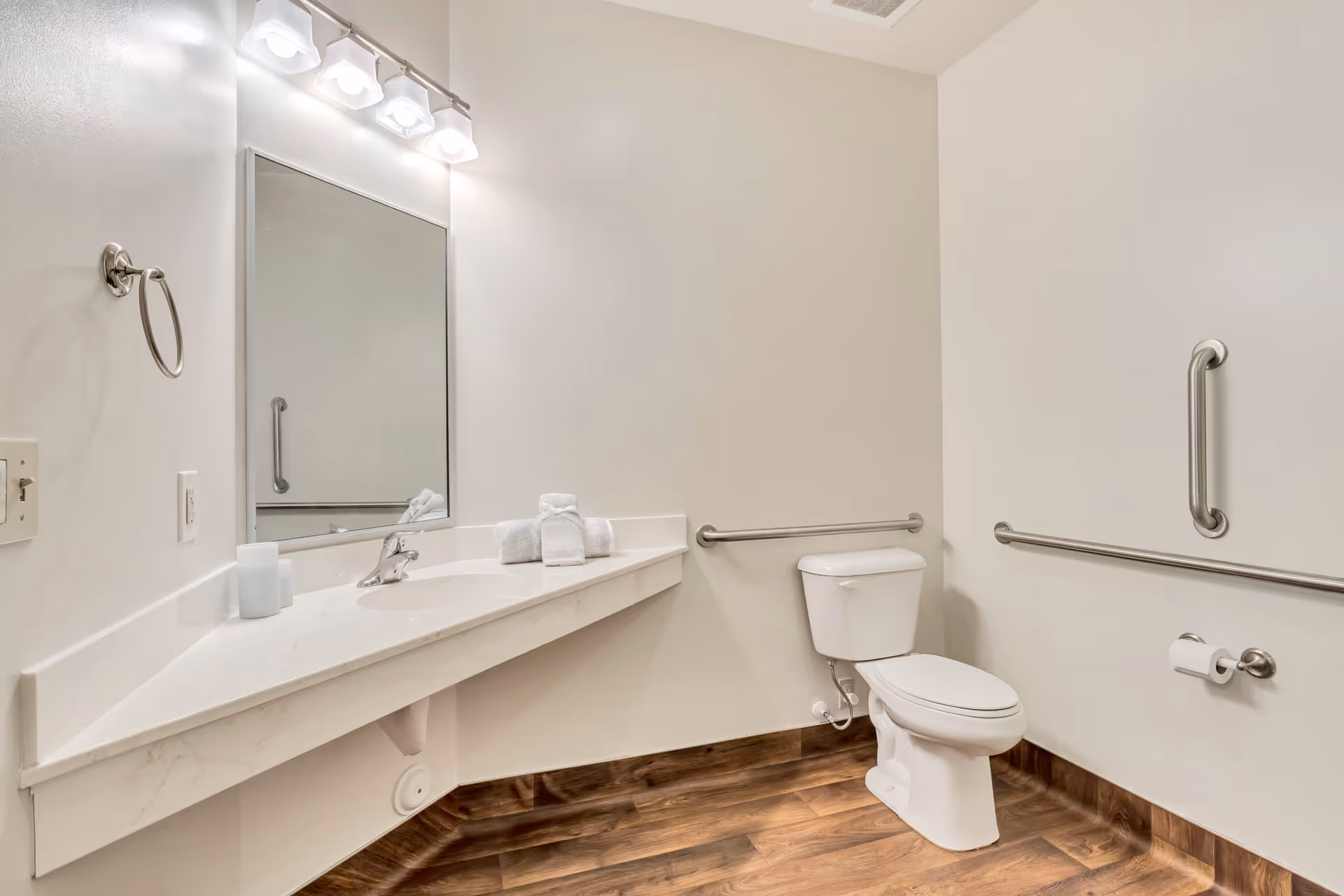 A clean, accessible bathroom with a white toilet, a large mirror above a white countertop with a sink, and three light fixtures above the mirror. The bathroom has grab bars on the walls near the toilet and a wooden floor.
