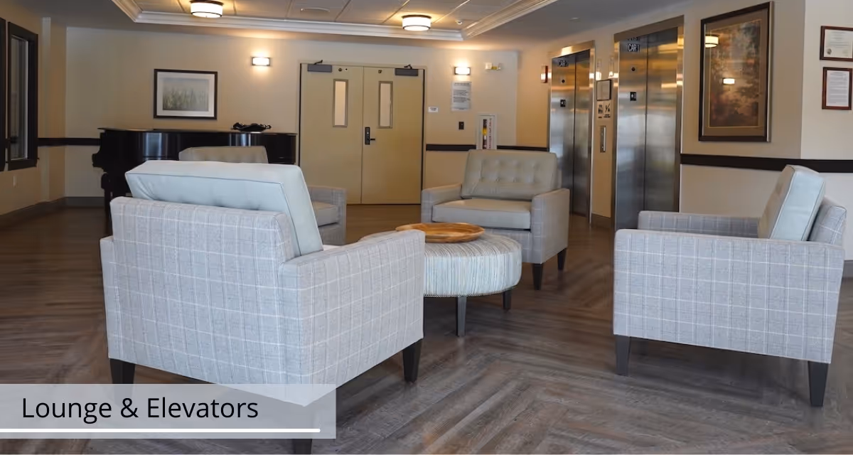 A senior living facility lounge with upholstered chairs arranged around a round ottoman and elevators in the background.