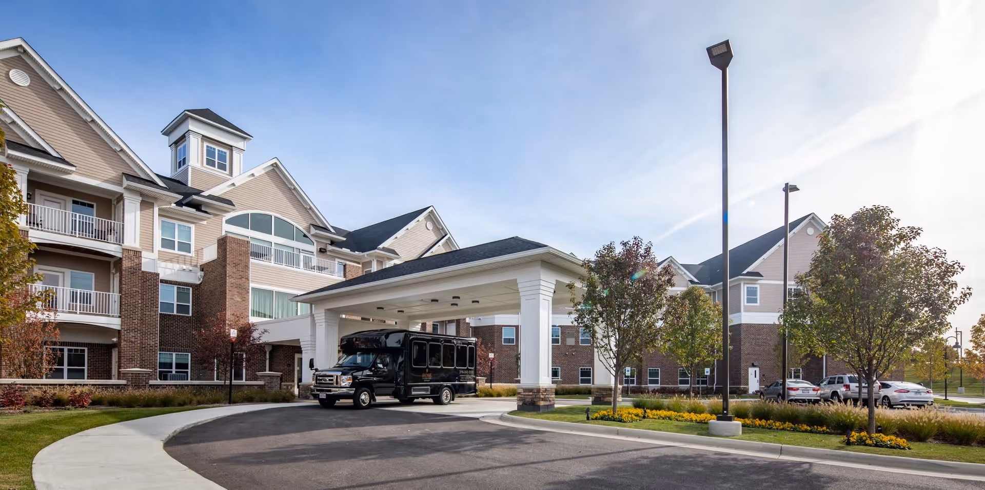 Exterior view of Rose Senior Living Beachwood, showing a multi-story building with balconies, a covered entrance with a black shuttle bus parked underneath, surrounded by trees and a parking area.