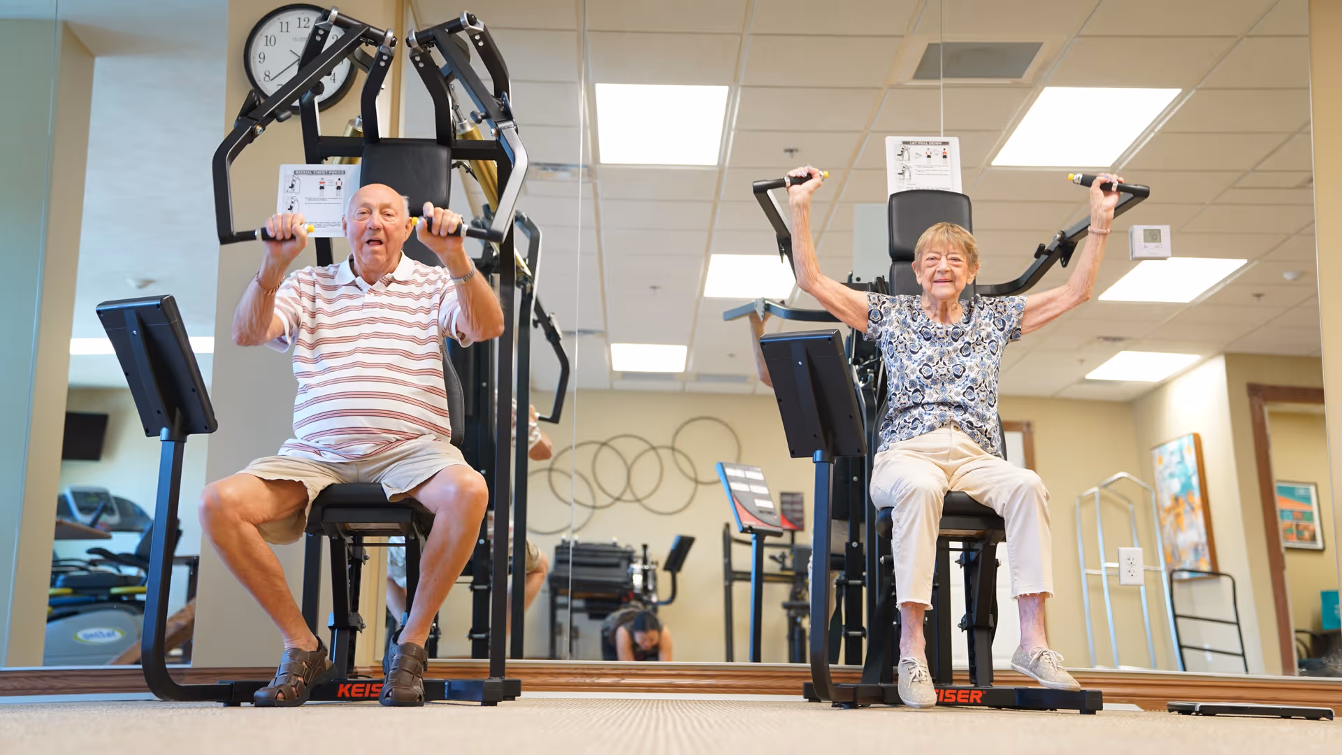 An elderly man and woman are seated on exercise machines in a fitness room, lifting weights with their arms raised. The room has a large mirror on the wall behind them, exercise equipment, and bright overhead lighting.