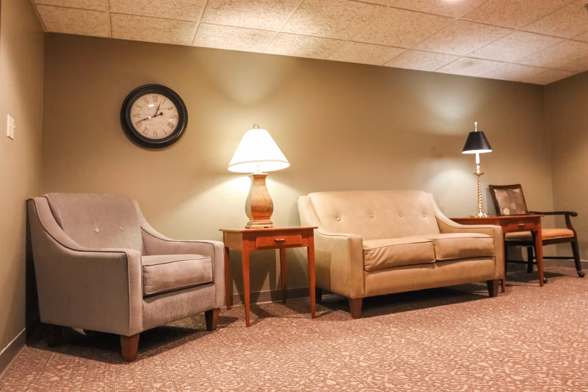 A cozy seating area with a beige sofa, a gray armchair, two wooden side tables each with a lamp, and a wooden chair with a cushion. A round wall clock with Roman numerals hangs on the wall above the armchair. The room has beige walls and patterned carpet flooring.