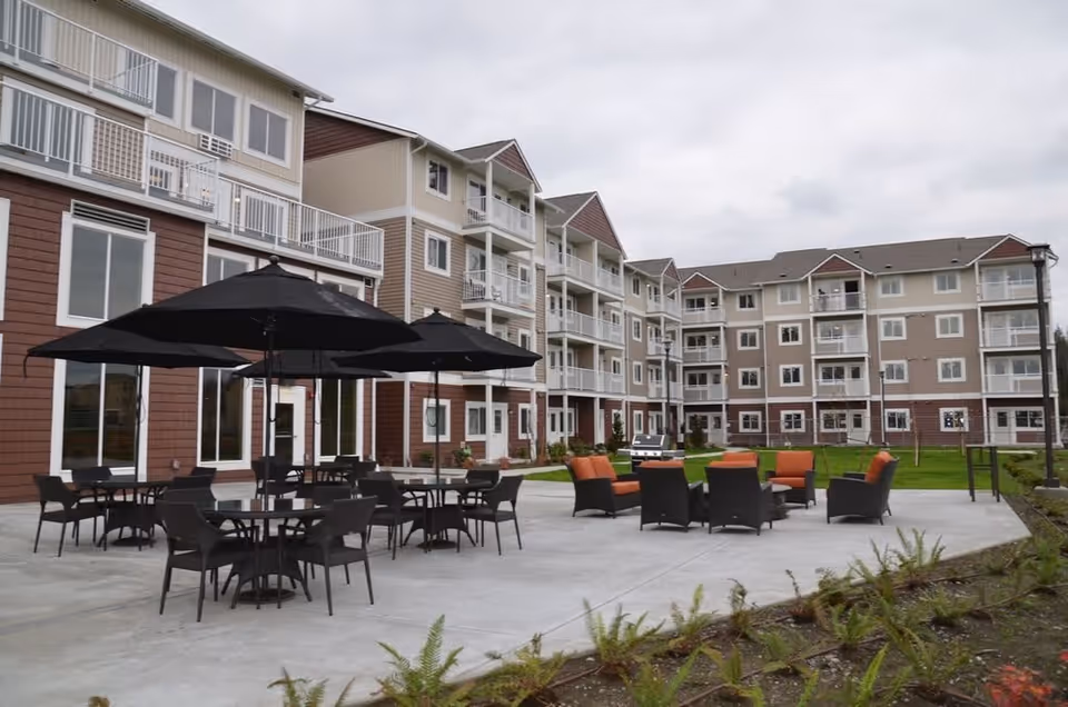 Outdoor patio area at Vintage at Lakewood featuring multiple black tables with black umbrellas and chairs, along with a seating area of cushioned chairs arranged around a fire pit. The patio is adjacent to a multi-story residential building with balconies and large windows under a cloudy sky.