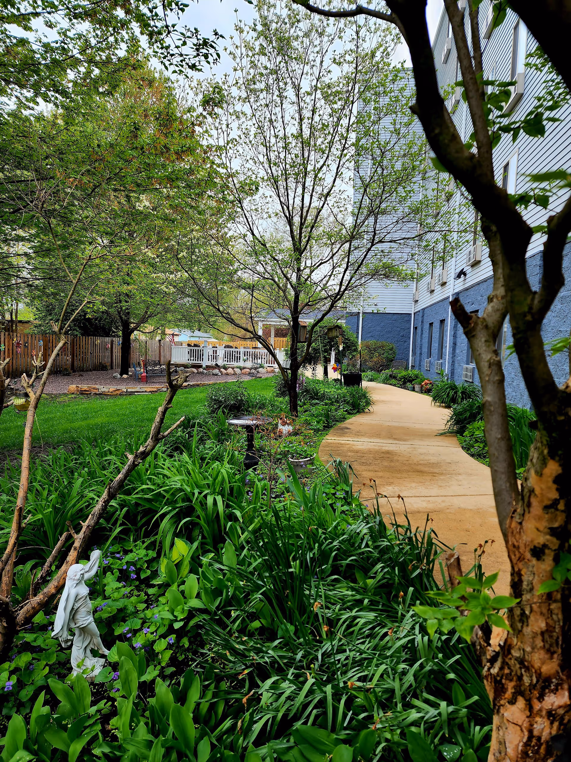 A landscaped outdoor garden area with a winding concrete pathway beside a building. The garden features lush green plants, small trees, a birdbath, and a white statue. A wooden fence and a white deck are visible in the background.