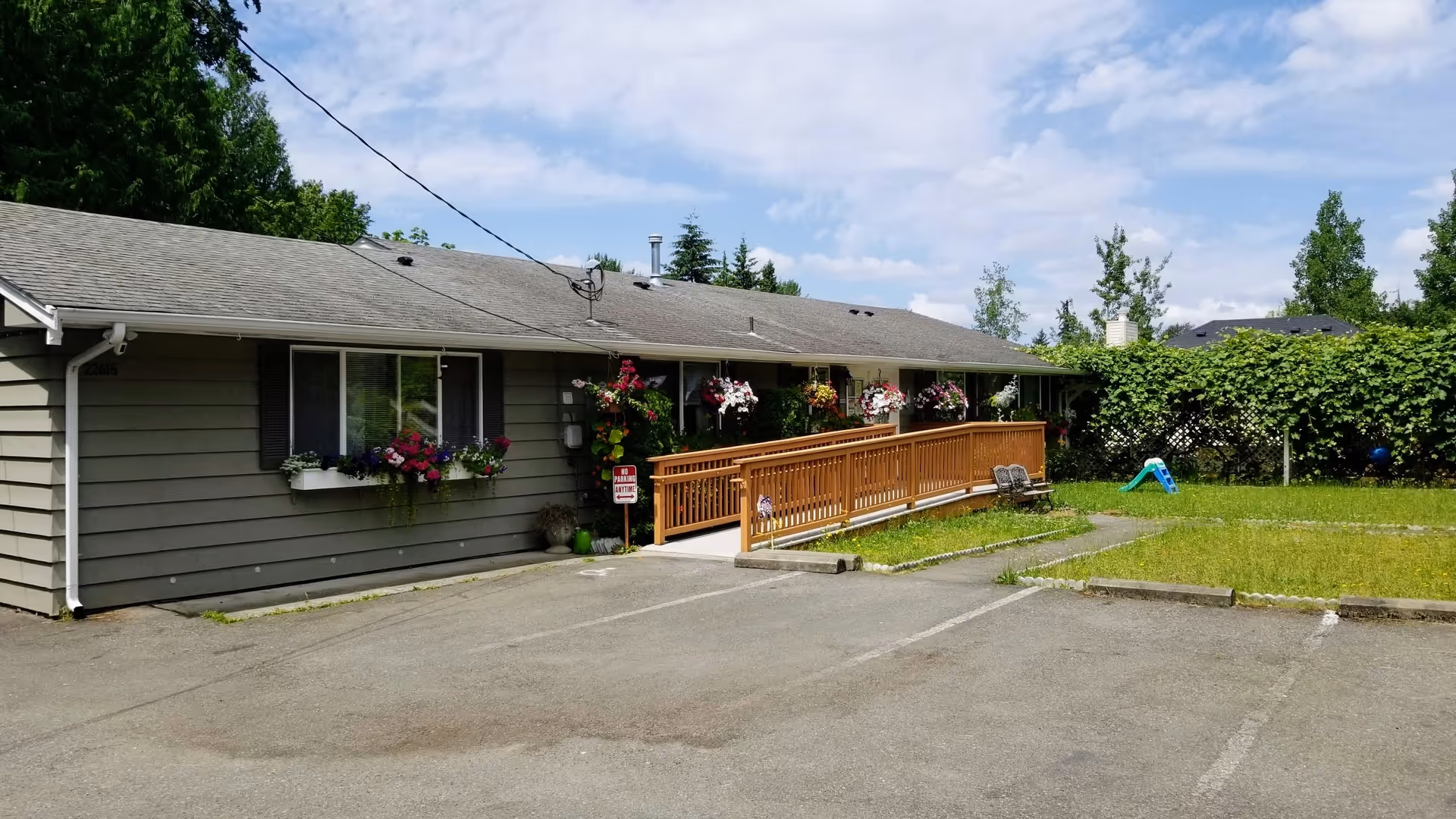 Single-story gray building with a wooden wheelchair ramp, flower boxes and a small parking area in front under a partly cloudy sky.